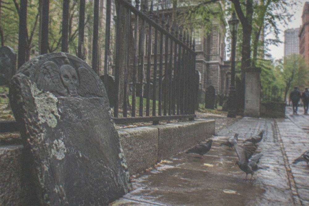A weathered colonial-era slate gravestone carved with a winged skull motif leaning in a fenced city cemetery