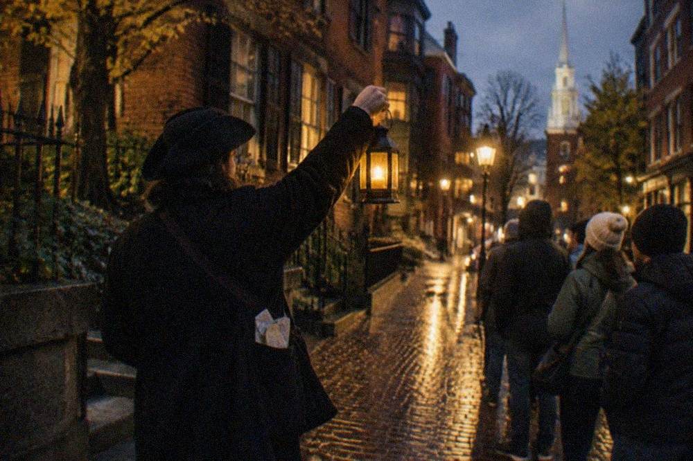 A tour guide holding a lantern while speaking to a small group on a dim Boston sidewalk at night.