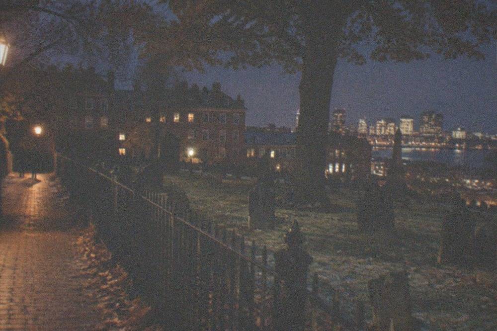 Gravestones and a wrought iron fence beside a lamplit path in an old Boston burying ground.