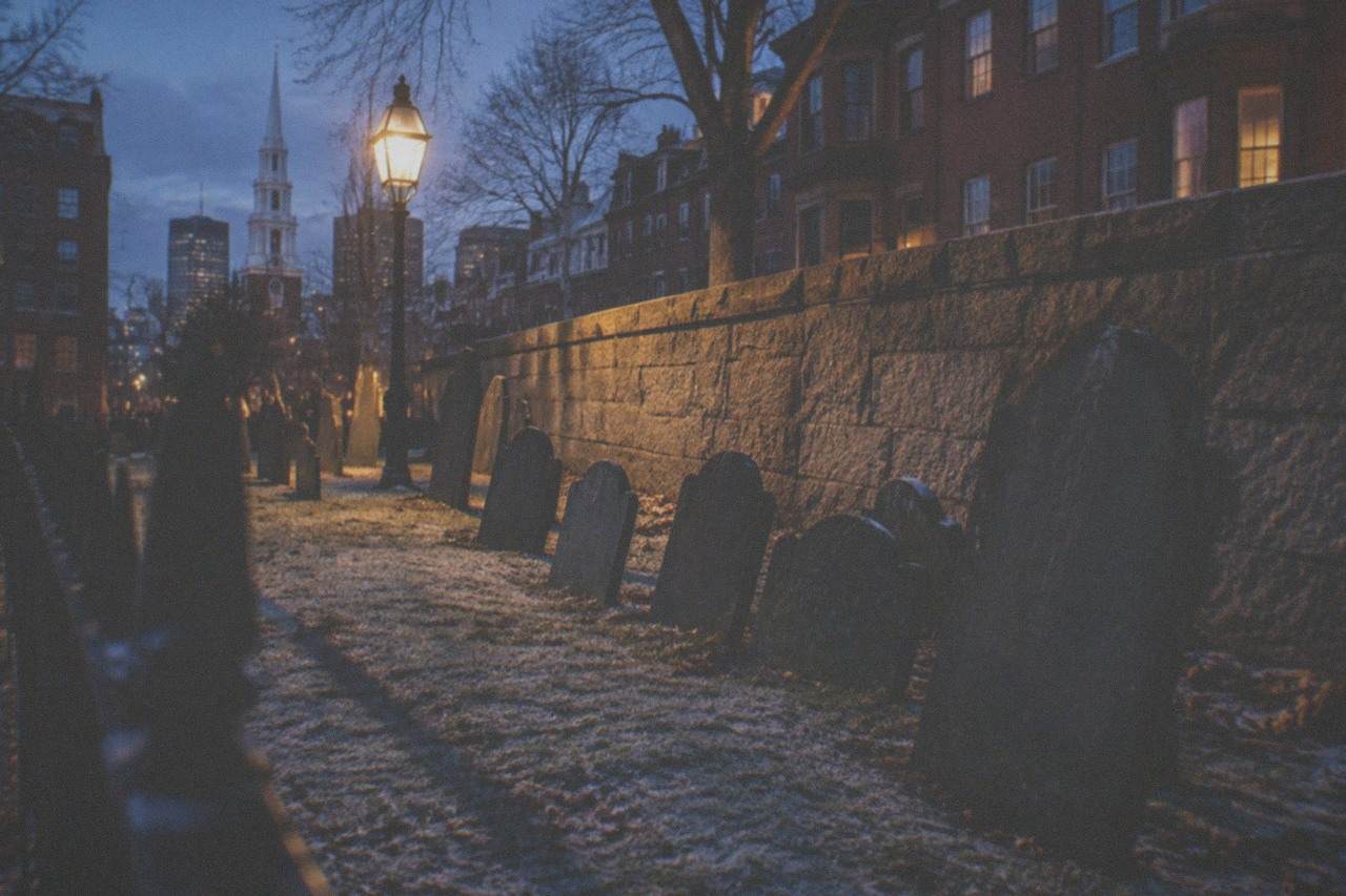 Lantern light glowing on old slate gravestones along a Boston burying ground fence at dusk.