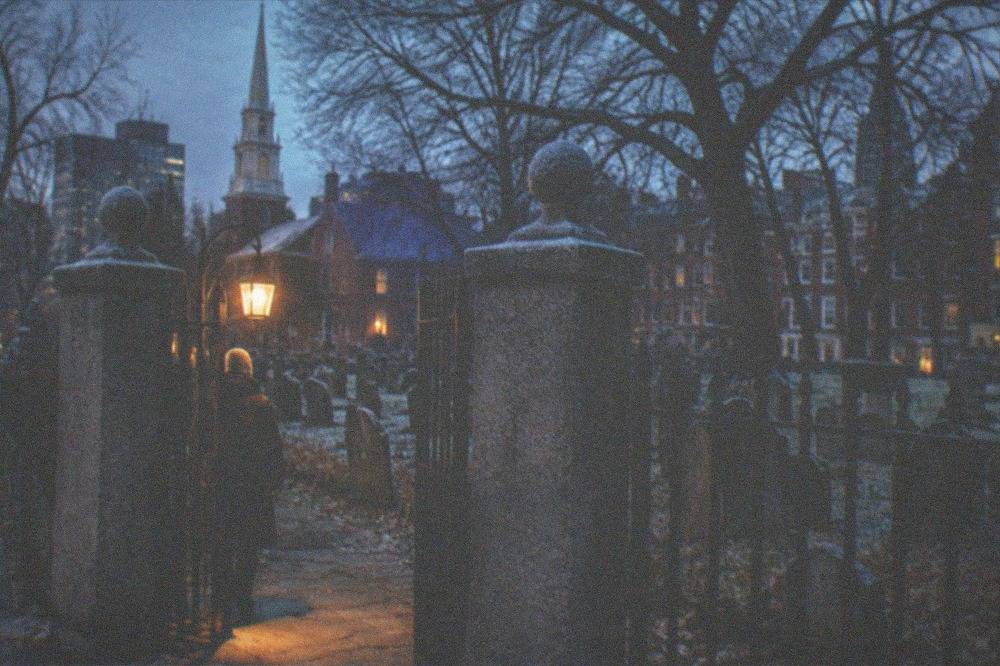 A lantern-lit colonial burying ground gate at dusk with iron fence and old gravestones beyond
