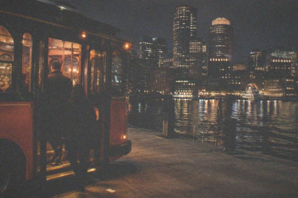 An orange and green vintage trolley stopped on a Boston street at night with the interior lit warmly.