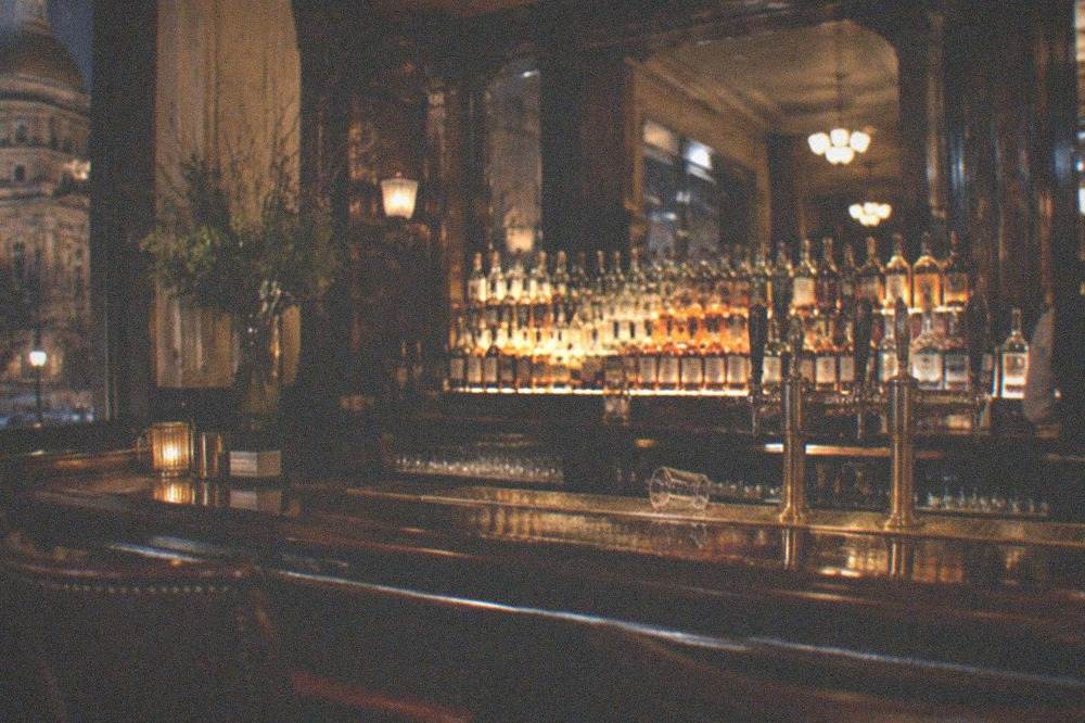 A dim historic hotel bar with dark wood, brass fixtures and a row of bottles backlit on shelves.