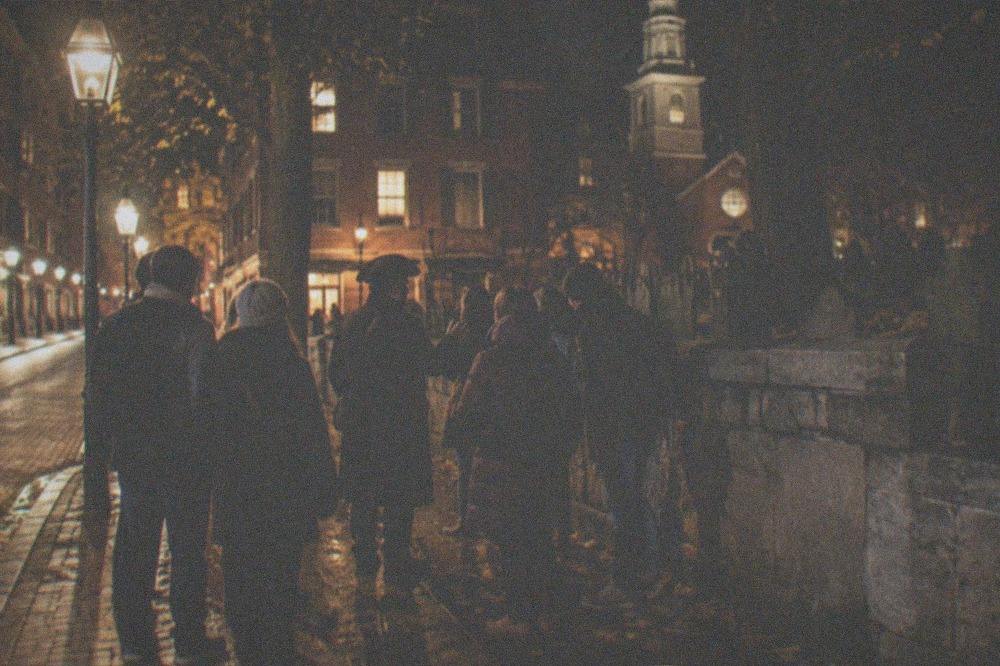 A Boston street corner at night with warm lantern light on brick facades and a small tour group gathered.