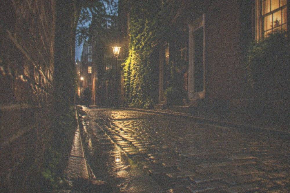 A narrow gaslit Boston alley at night with brick walls and a distant streetlamp