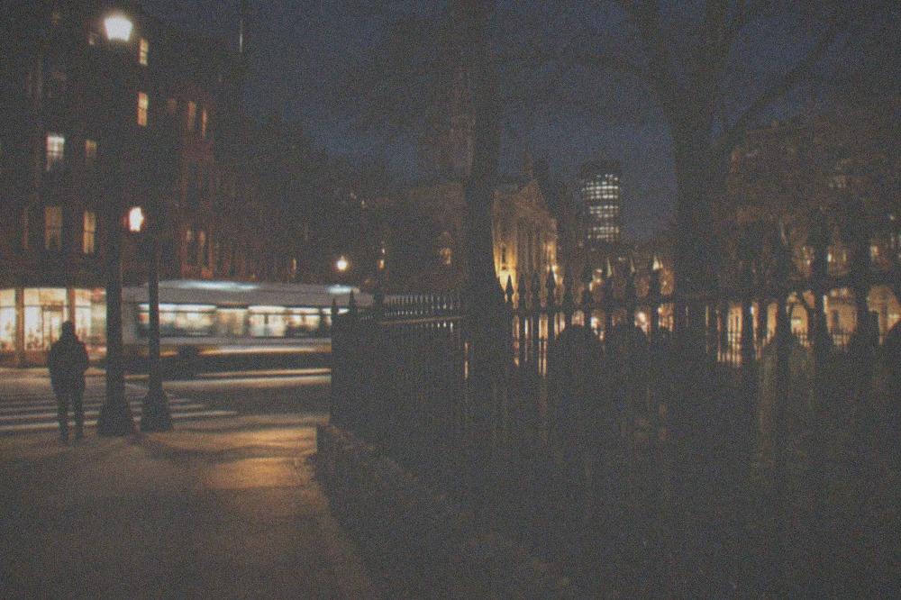 A nighttime view down Tremont Street with cemetery fence silhouettes and warm shop lights