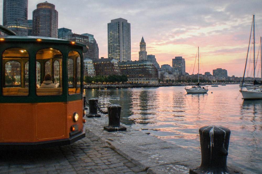 A vintage-style orange and green trolley parked on a Boston waterfront street at dusk