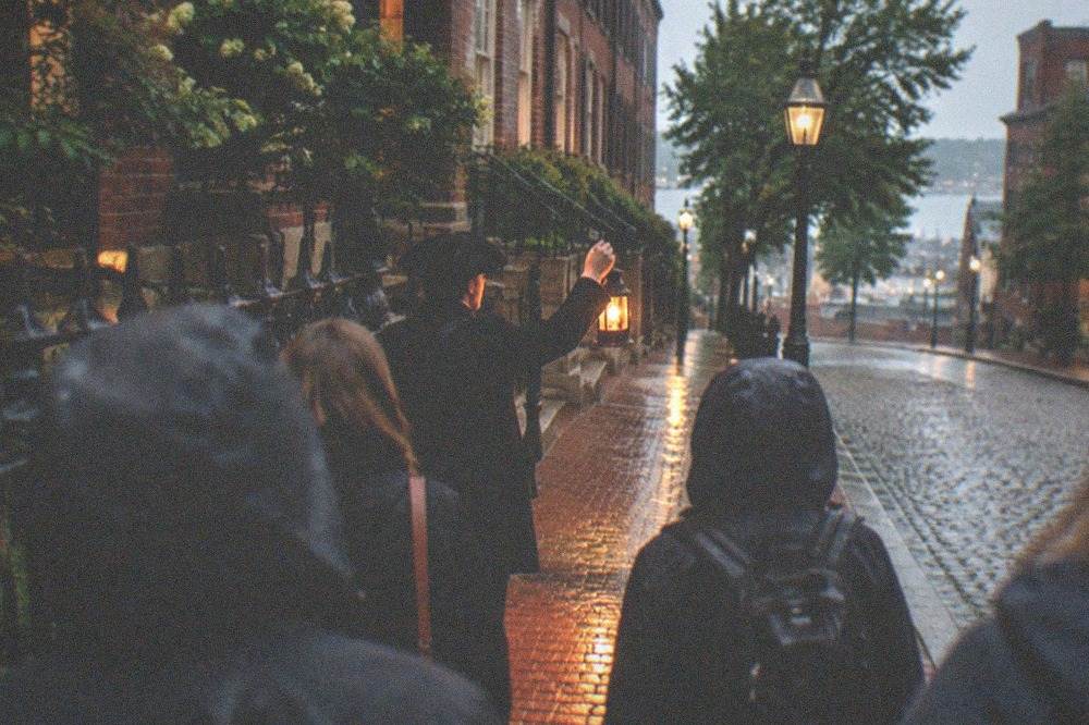 A guide holding a lantern and speaking to a small evening tour group on a Boston sidewalk