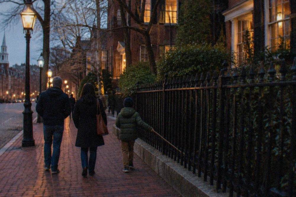 A family walking a lamp-lit brick path past iron fencing in downtown Boston at dusk