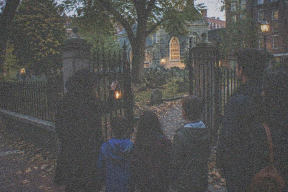 A guide holding a lantern addressing a small gathered group on a sidewalk beside an old cemetery gate