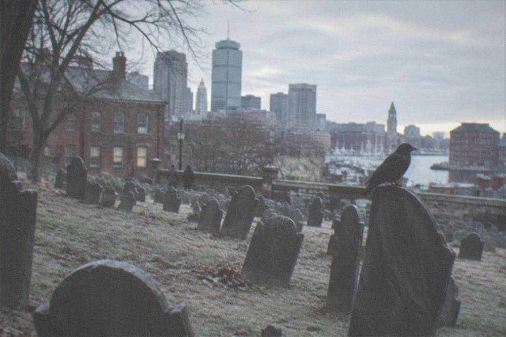 Rows of weathered slate gravestones inside a colonial burying ground bordered by city buildings