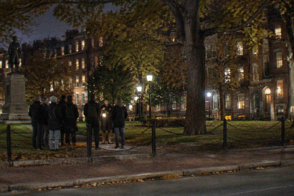 Crowd of adults gathered around a tour guide outside a lantern-lit Boston bar at night