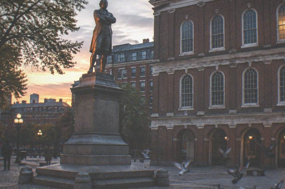 Bronze statue of a colonial-era figure beside a historic Boston building at dusk