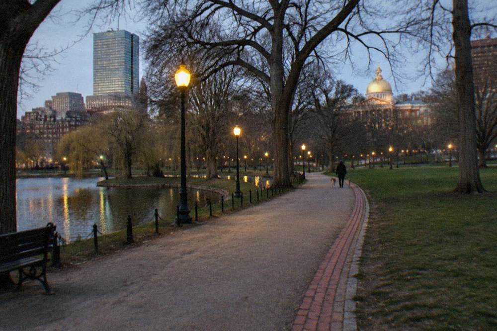 A lamp-lit walking path curving through Boston Common at twilight with bare trees