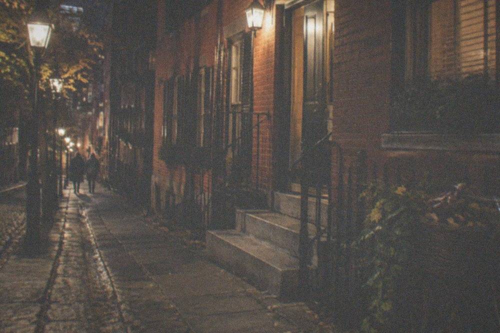 A narrow gaslit-style street in old Boston at night with brick rowhouses and worn sidewalks