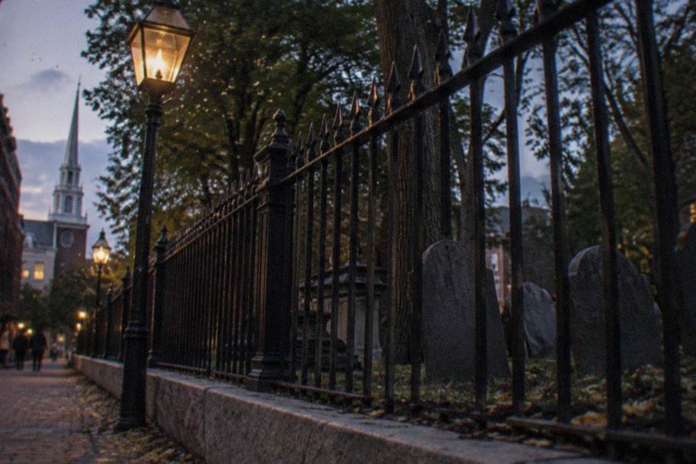 A colonial-era slate gravestone yard enclosed by an iron fence beside a gas-lit Boston street at twilight.
