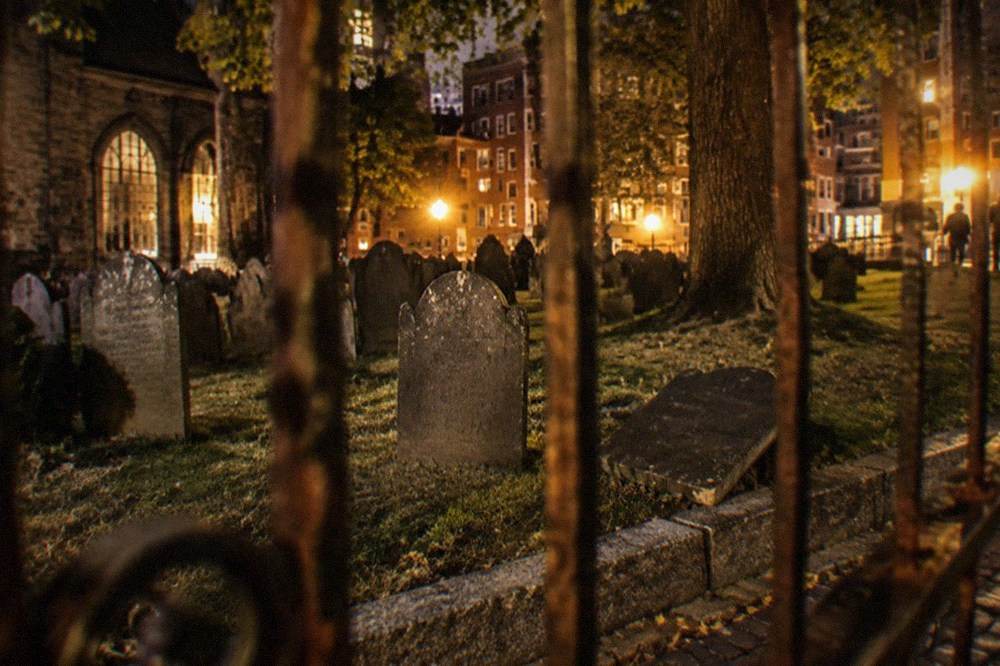 Rows of weathered slate gravestones inside a fenced colonial burying ground lit by street lamps.