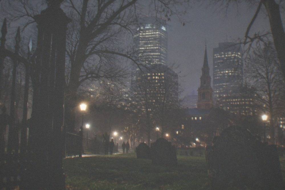 An old stone burial ground at night with a wrought iron gate and city office towers glowing behind it.