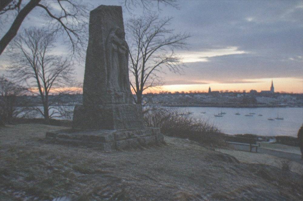 An old stone monument surrounded by bare trees near a harbor at dusk