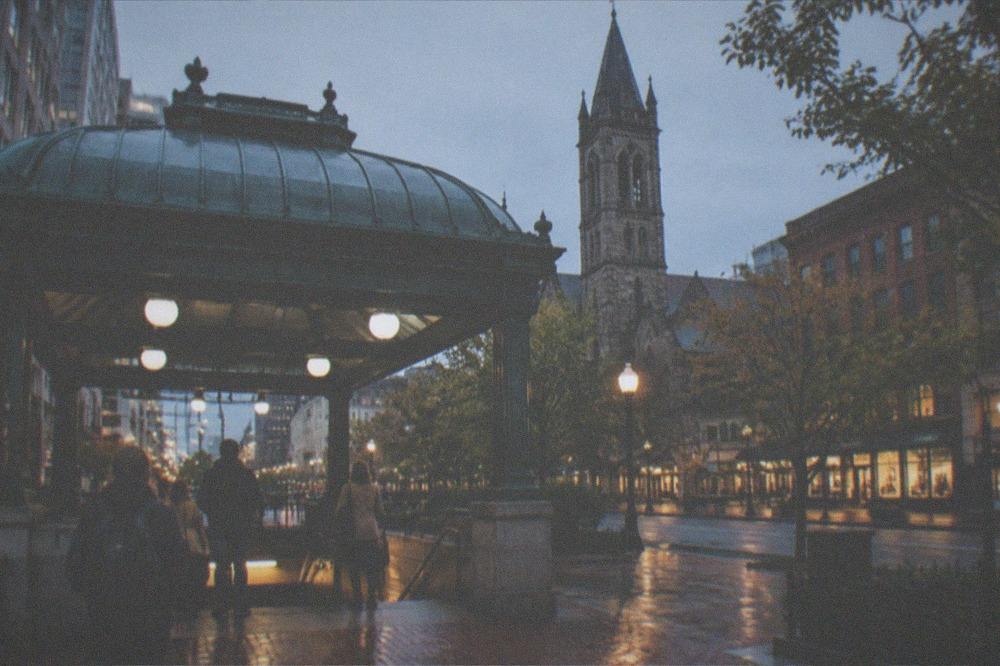 An evening meeting spot at a Boston street corner with lit storefronts and a historic church behind